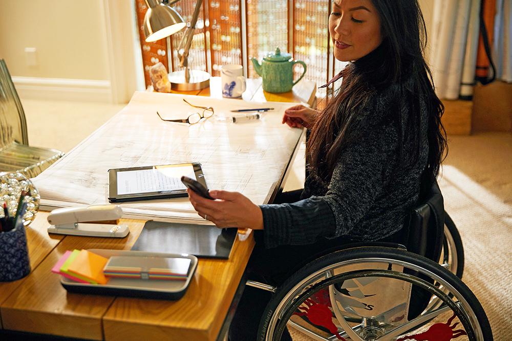 A female wheelchair sits at a desk, looking at her phone