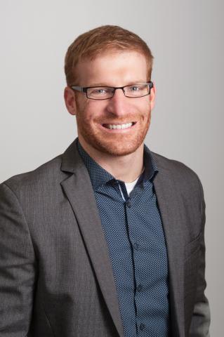Head shot of Jeff Dusek with red hair and black glasses wearing a blue collared shirt and a grey blazer.